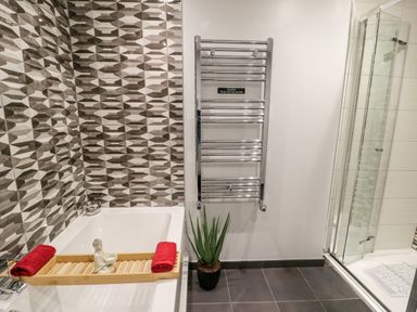 A bathroom with a bathtub with a wooden tray and red towels a chrome towel rail a potted plant and a glass shower enclosure at The Chalet in Benllech