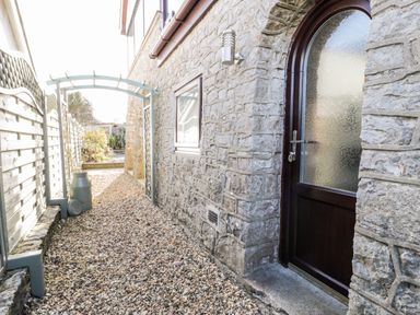 A stone wall with an arched wooden door and frosted window beside a narrow gravel path and a white wooden fence with an archway over the path at The Chalet in Benllech