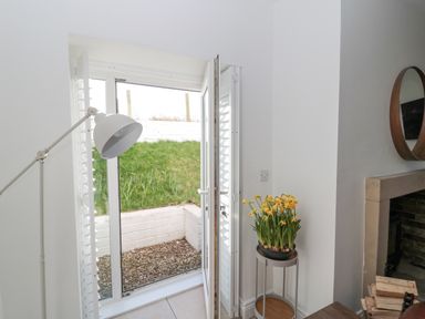 A room with an open glass door showing a small outdoor area with grass and white brick wall and a plant with yellow flowers on a stand next to a fireplace at Little Haven in Bamburgh