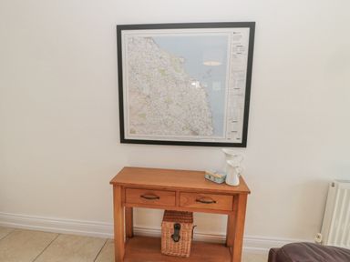 A wooden console table with two drawers a wicker basket underneath and two white pitchers on top below a framed map on a white wall
