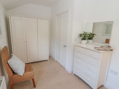 A room with a white wardrobe a white chest of drawers with a mirror and a wicker chair with a cushion at Little Haven in Bamburgh