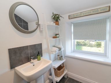 A bathroom with a wall-mounted sink, round mirror, white ladder shelf with towels and decor, and a window with a shade at Little Haven in Bamburgh