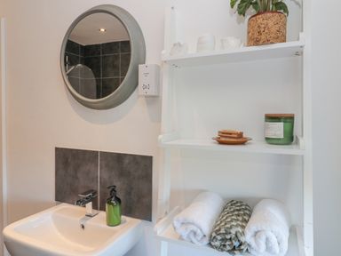 A bathroom sink with a green soap dispenser a round mirror and a white shelf holding rolled towels a plant a brush and a green candle at Little Haven Bamburgh