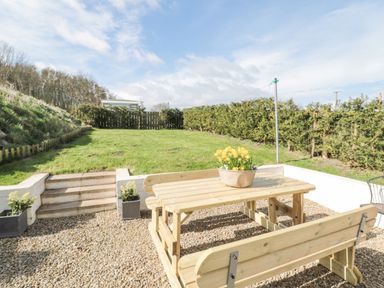 A garden with a wooden picnic table and benches on a gravel area and grass with hedges at Little Haven in Bamburgh