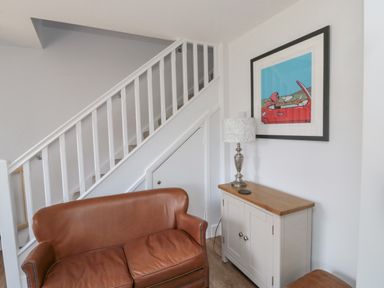 A brown leather sofa under white stairs next to a small wooden cabinet with a lamp and framed art on the wall at 3 St Cuthberts Garth in Bamburgh
