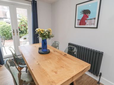 A dining area with a wooden table and four transparent chairs with a vase of flowers on the table and framed artwork on the wall at 3 St Cuthberts Garth in Bamburgh