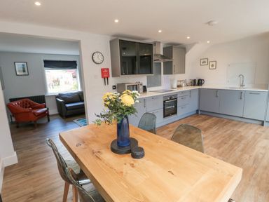 A kitchen with wooden dining table and flowers in a vase next to a living room with leather chairs at 3 St Cuthberts Garth in Bamburgh