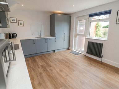 A kitchen with grey cabinets wooden floor a sink a microwave kettle toaster radiator and a door with window at 3 St Cuthberts Garth in Bamburgh