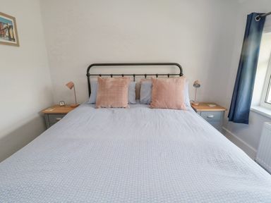 A bedroom with a bed with blue bedding and pink pillows two bedside tables with lamps and a window with blue curtains at 3 St Cuthberts Garth in Bamburgh