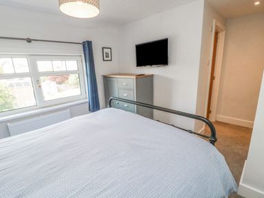A bedroom with a bed a window with blue curtains a dresser and a wall-mounted TV at 3 St Cuthberts Garth in Bamburgh