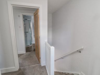 A hallway with carpeted stairs and an open wooden door leading to a bathroom with a curved glass shower enclosure at 3 St Cuthberts Garth in Bamburgh