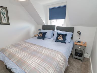 A bedroom with a double bed two pillows with bee designs a plaid blanket two bedside tables with lamps and a window with a blue blind at 3 St Cuthberts Garth in Bamburgh