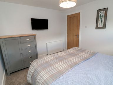 A bedroom with a bed covered with a blanket and duvet a grey cabinet on the left a wall-mounted TV a wooden door and a framed picture on the wall at 3 St Cuthberts Garth in Bamburgh