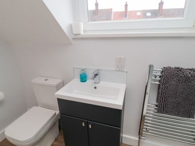 A bathroom with a toilet a sink with cabinet a hand soap dispenser a radiator with a towel and a window above at 3 St Cuthberts Garth in Bamburgh