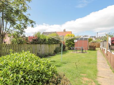 A backyard with a wooden fence green grass a rotary clothesline a small table with chairs and garden sheds at 3 St Cuthberts Garth in Bamburgh