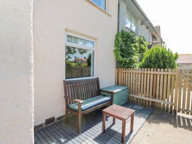 An outdoor seating area with a wooden bench cushion small wooden table next to a beige wall and wooden fence at 3 St Cuthberts Garth in Bamburgh