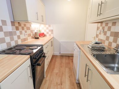 A galley kitchen with wooden floors white cabinets a black electric stove a stainless steel sink a kettle and a red toaster at Windermere Crescent in Windermere