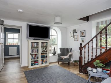 A living room with a mounted TV above a cabinet with DVDs and board games a grey armchair under framed pictures and a wooden staircase with a window and plant at Bethania in Llangoed near Beaumaris