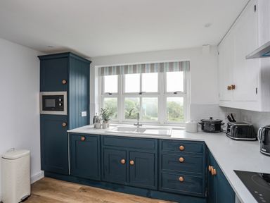 A kitchen with blue lower cabinets white upper cabinets appliances and a window with a striped blind at Bethania in Llangoed near Beaumaris