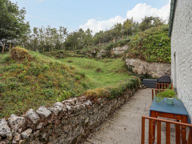 A narrow outdoor patio next to a house with a table, chairs, a barbecue, and a stone wall with grass and trees beyond at Bethania in Llangoed near Beaumaris