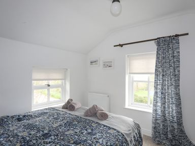 A bedroom with a bed covered in patterned bedding and three rolled towels near two windows with blinds and one curtain at Bethania in Llangoed near Beaumaris