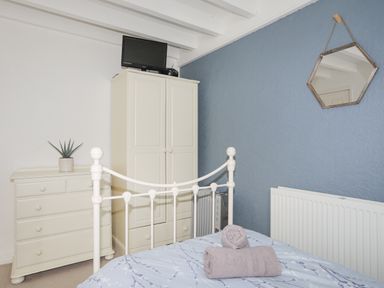 A bedroom with a white dresser, wardrobe, TV, bed with folded towels, and hexagonal mirror on blue wall at Bethania in Llangoed near Beaumaris