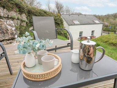 An outdoor seating area with a metal table holding a tray with mugs and a vase and a metal teapot with a small jug and a chair on a wooden deck at Bethania in Llangoed near Beaumaris