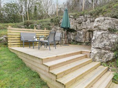 An outdoor wooden deck with chairs a table and a closed umbrella near rocky terrain at Bethania in Llangoed near Beaumaris