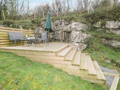 A wooden deck with steps leading up to a seating area with chairs a table and an umbrella on grass near rock formations at Bethania in Llangoed near Beaumaris
