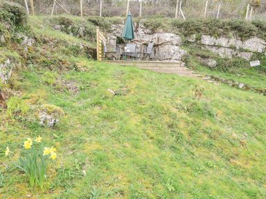 An outdoor seating area with table and umbrella on a wooden platform surrounded by grassy rocks and yellow flowers at Bethania in Llangoed near Beaumaris