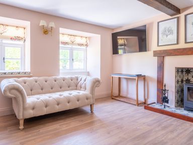 A living room with a beige tufted sofa two windows a wall-mounted TV a wooden console table and a fireplace at Cunsey Mill House in Cunsey near Hawkshead