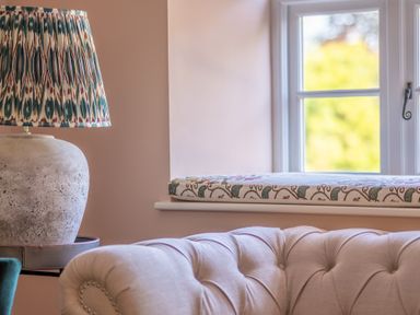 A room corner with a patterned lampshade on a stone base lamp a cushioned beige sofa and a window with a floral cushion on the sill at Cunsey Mill House Cunsey near Hawkshead