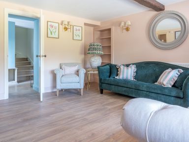 A living room with a green sofa, a beige armchair, a side table with a lamp, wall sconces, framed artwork, a round mirror, and an open door showing stairs at Cunsey Mill House Cunsey near Hawkshead