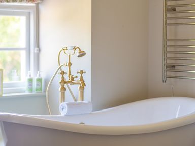 A white freestanding bathtub with a rolled towel on the edge next to a gold faucet and window at Cunsey Mill House Cunsey near Hawkshead