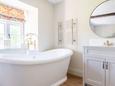 A bathroom with a freestanding bathtub a window with patterned curtains a towel radiator a round mirror and a vanity unit at Cunsey Mill House Cunsey near Hawkshead