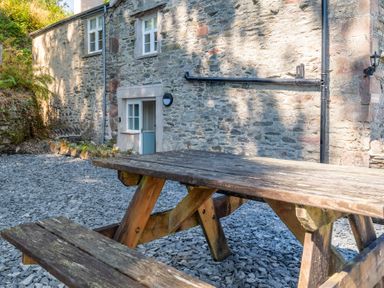 An outdoor wooden picnic table on a gravel ground next to a stone building with windows and a door at Cunsey Mill House in Cunsey near Hawkshead