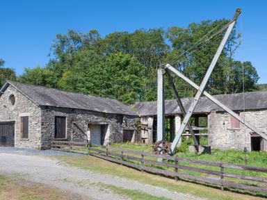 Stone buildings with timber frames and machinery behind a wooden fence surrounded by greenery at Cunsey Mill House in Cunsey near Hawkshead