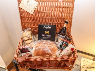 A wicker basket with food items including bread pasta biscuits beer and jam at The Cheese Room in Stretton-On-Fosse