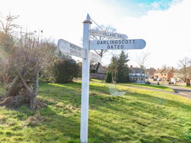 A grassy roadside area with a wooden signpost showing directions to local places and houses in the background at The Cheese Room in Stretton-On-Fosse