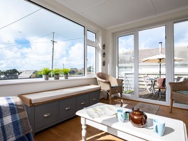 A sunroom with wicker chairs a bench with cushions a white coffee table and large windows overlooking an outdoor patio at Croft Elms in Trebetherick near Polzeath