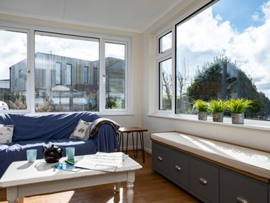 A sitting area with a blue covered sofa two cushions a white coffee table with a teapot two mugs and maps a bench with storage and potted plants on the windowsill at Croft Elms in Trebetherick near Polzeath