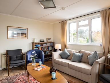 A living room with a beige sofa and blue pillows a wooden coffee table with cups and a vase two armchairs a bookshelf and a large window at Croft Elms in Trebetherick near Polzeath