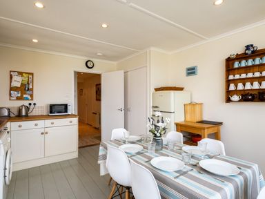 A kitchen with a dining table set with plates glasses and cutlery a fridge a wooden shelf holding cups and teapots and a microwave on a counter at Croft Elms in Trebetherick near Polzeath