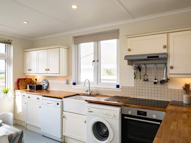 A kitchen with white cabinets wooden countertops a dishwasher washing machine oven and a window at Croft Elms in Trebetherick near Polzeath