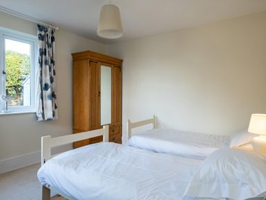 A bedroom with two single beds white bedding a wooden wardrobe a window with curtains and a bedside lamp at Croft Elms in Trebetherick near Polzeath
