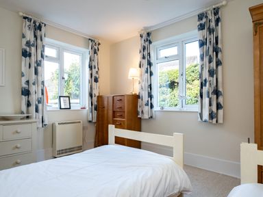 A bedroom with two beds a wooden wardrobe a chest of drawers and windows with patterned curtains at Croft Elms in Trebetherick near Polzeath