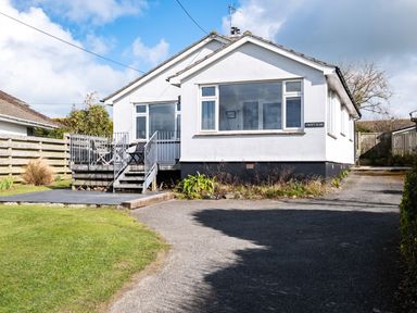 A white bungalow with a front porch and outdoor chairs and table surrounded by grass and driveway at Croft Elms in Trebetherick near Polzeath