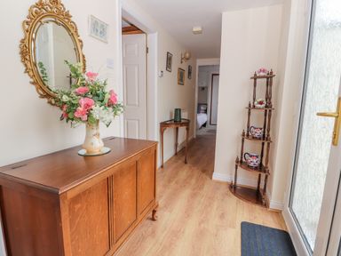A hallway with wooden flooring a wooden cabinet with a flower vase a decorative wall mirror a wooden corner shelf with ceramic items and a side table at Bryn Offa cottage in Holywell