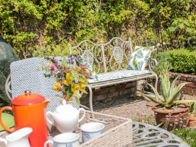 A garden seating area with a metal bench and cushions surrounded by plants and a table with tea set at Bryn Offa cottage in Holywell