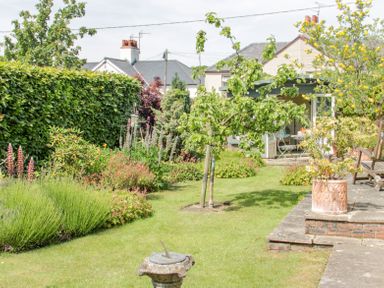 A garden with a variety of plants and flowers a small tree on a lawn a sundial on a pedestal a patio with potted plants a wooden bench and a small garden building with outdoor furniture at Bryn Offa cottage in Holywell